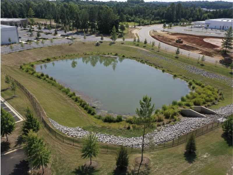 Aerial view of a man-made pond surrounded by grassy embankments and stone borders in a park-like area with trees and roads nearby
